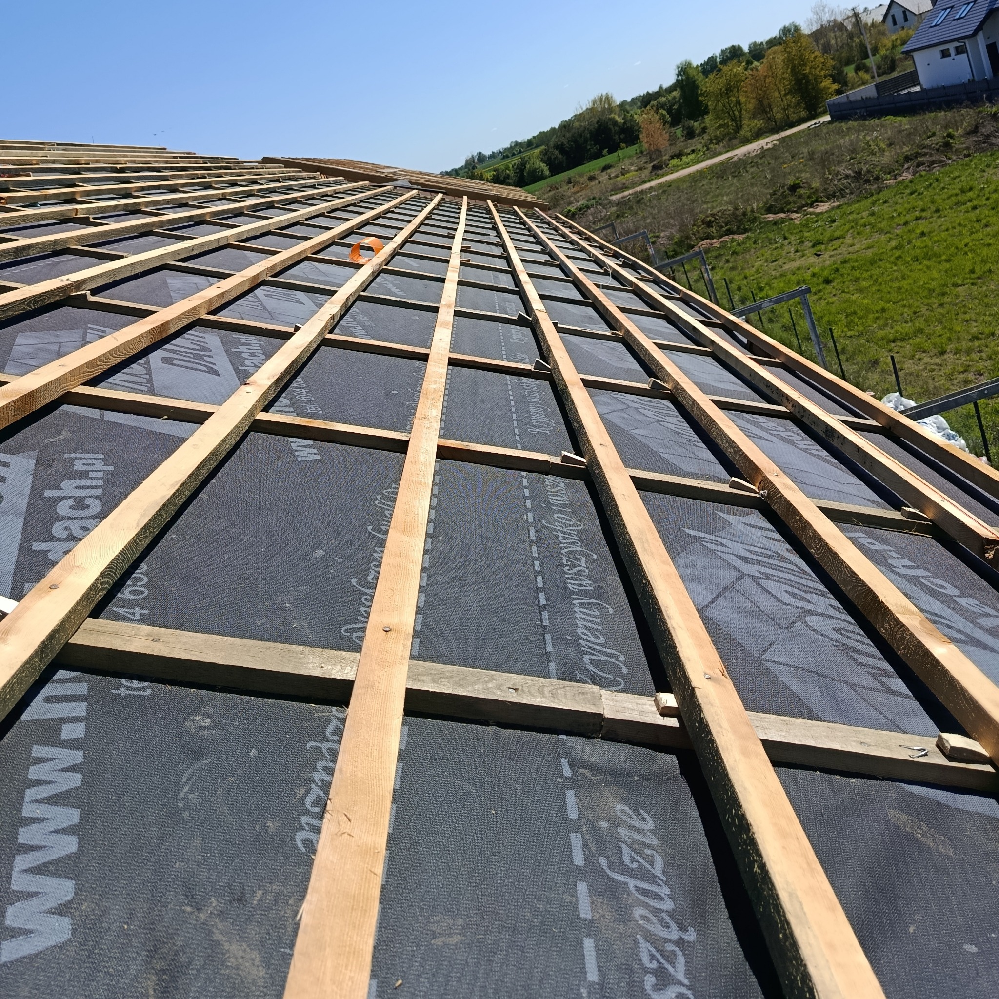 Konstrukcja dachu z drewnianych łat na czarnej membranie dachowej, widok z dołu pod kątem. W tle zieleń i budynki.