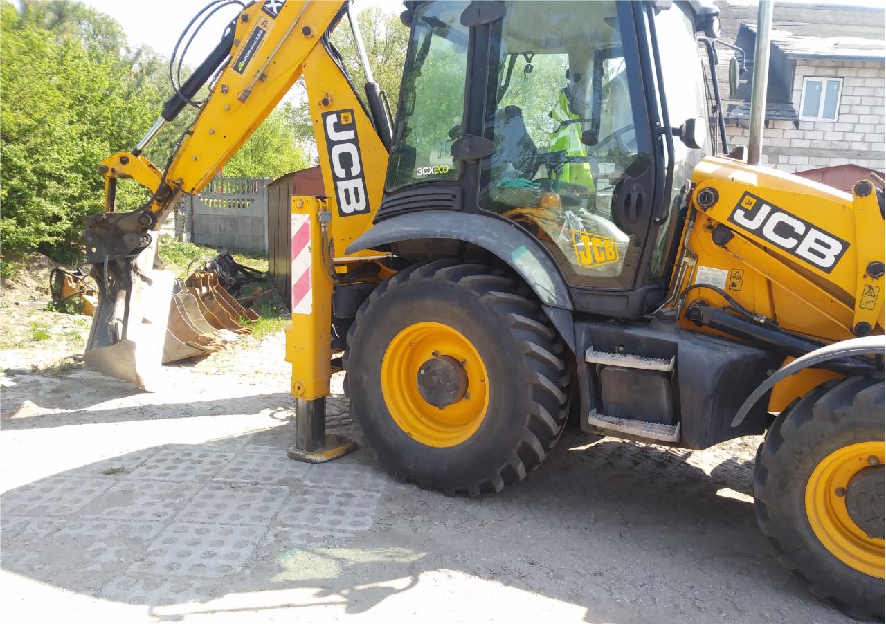 Żółto-czarna koparko-ładowarka JCB z opuszczoną stopą stabilizującą na betonowej kostce, w tle widoczne dodatkowe łyżki i zieleń.