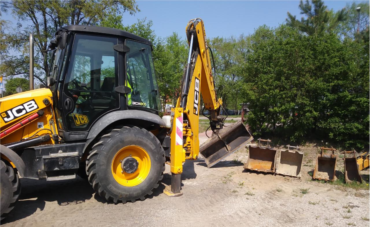 Żółto-czarna koparko-ładowarka JCB stoi na ziemi, z wysuniętą łyżką i kilkoma dodatkowymi łyżkami obok, drzewa w tle.