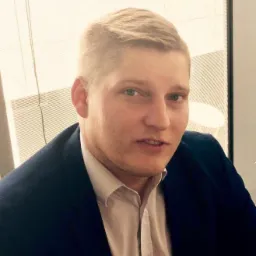 Portret młodego mężczyzny w ciemnej marynarce i jasnej koszuli, z kr&oacute;tkimi blond włosami, stojącego na tle biurowego okna z żaluzjami.