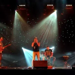 Występ na scenie: czterech muzyków w świetle reflektorów, tło z efektem gwiazd, jeden wokalista śpiewa do mikrofonu, dwaj gitarzyści grają, jeden siedzi na krześle, czwarty gra na perkusji.