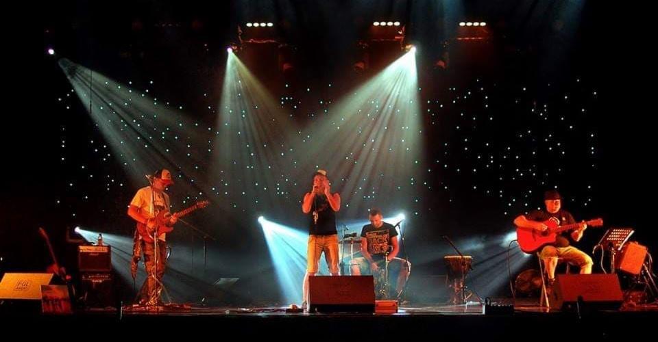 Występ na scenie: czterech muzyków w świetle reflektorów, tło z efektem gwiazd, jeden wokalista śpiewa do mikrofonu, dwaj gitarzyści grają, jeden siedzi na krześle, czwarty gra na perkusji.