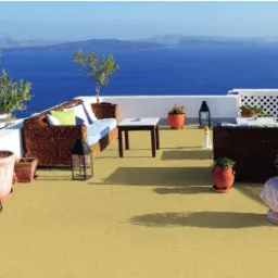 Umeblowany balkon z widokiem na morze, dwie sofy z wikliny z jasnymi poduszkami, stolik kawowy, liczne donice z roślinami, lampiony dekoracyjne, podłoga wyłożona wykładziną w kolorze piaskowym.