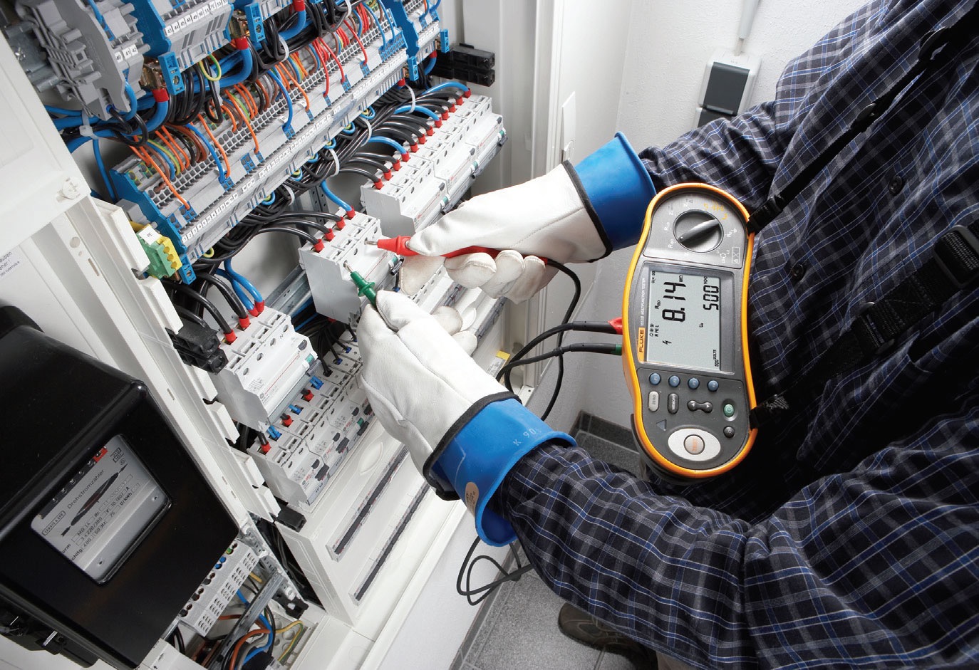 Elektryk w rękawicach ochronnych dokonuje pomiarów w rozdzielnicy elektrycznej za pomocą miernika Fluke, widoczne kable i bezpieczniki.
