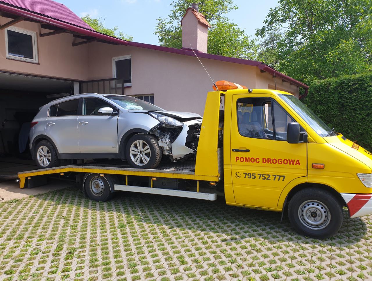 Żółty pojazd pomocy drogowej z uszkodzonym srebrnym SUV-em na platformie, widoczne uszkodzenia przodu pojazdu, dom z czerwonym dachem w tle.