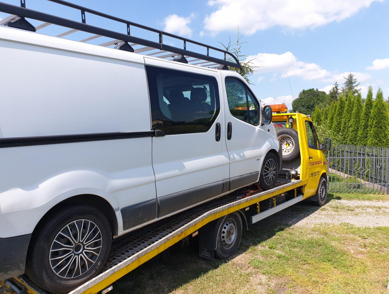 Żółty pojazd pomocy drogowej przewozi białego vana z bagażnikiem dachowym, widoczne numery telefonu na karoserii, w tle zielone tuje i błękitne niebo.