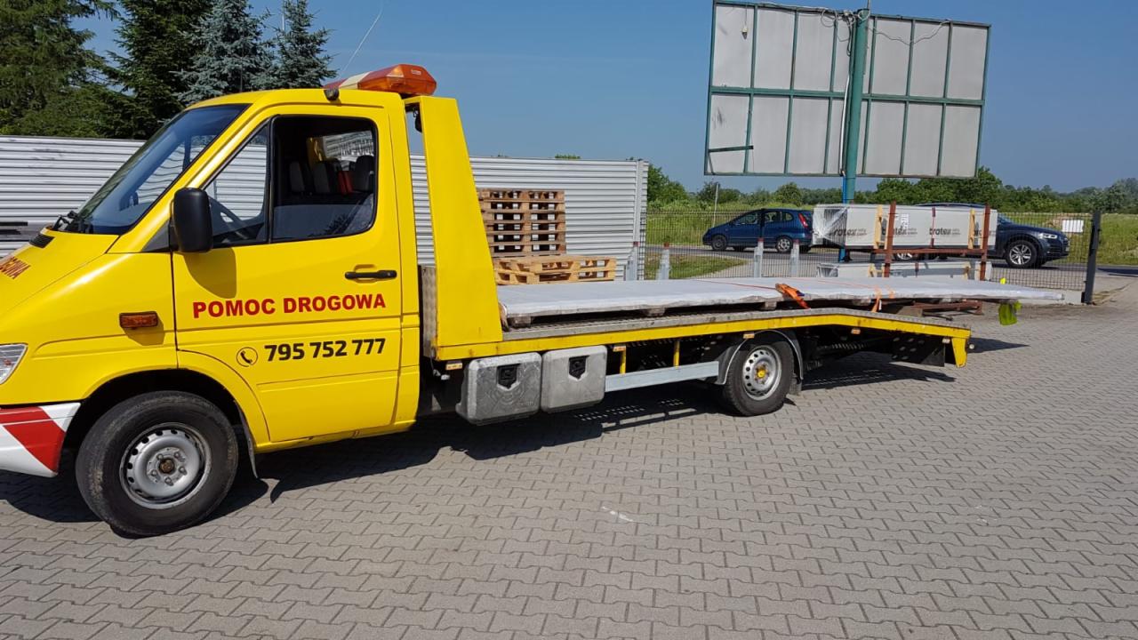 Żółta laweta z napisem 'Pomoc Drogowa' i numerem telefonu, przewożąca płyty betonowe na tle parkingu i zieleni.