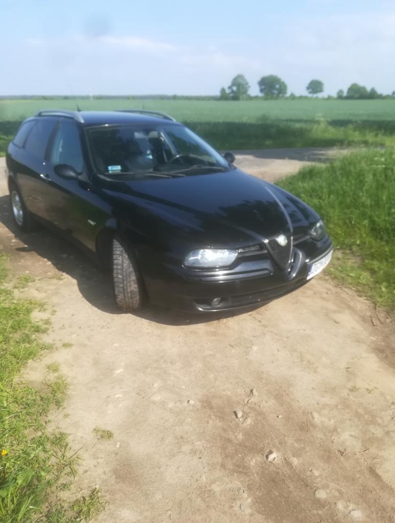 Czarna Alfa Romeo 156 Sportwagon na polnej drodze z zielonym polem w tle, widoczne relingi dachowe.