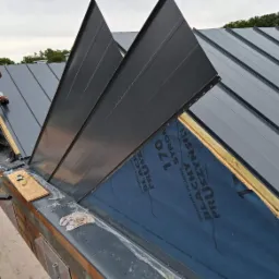 Prace dekarskie: montaż paneli z blachy na dachu, widoczna membrana dachowa, dekarz mocuje element, narzędzia leżą na murze.