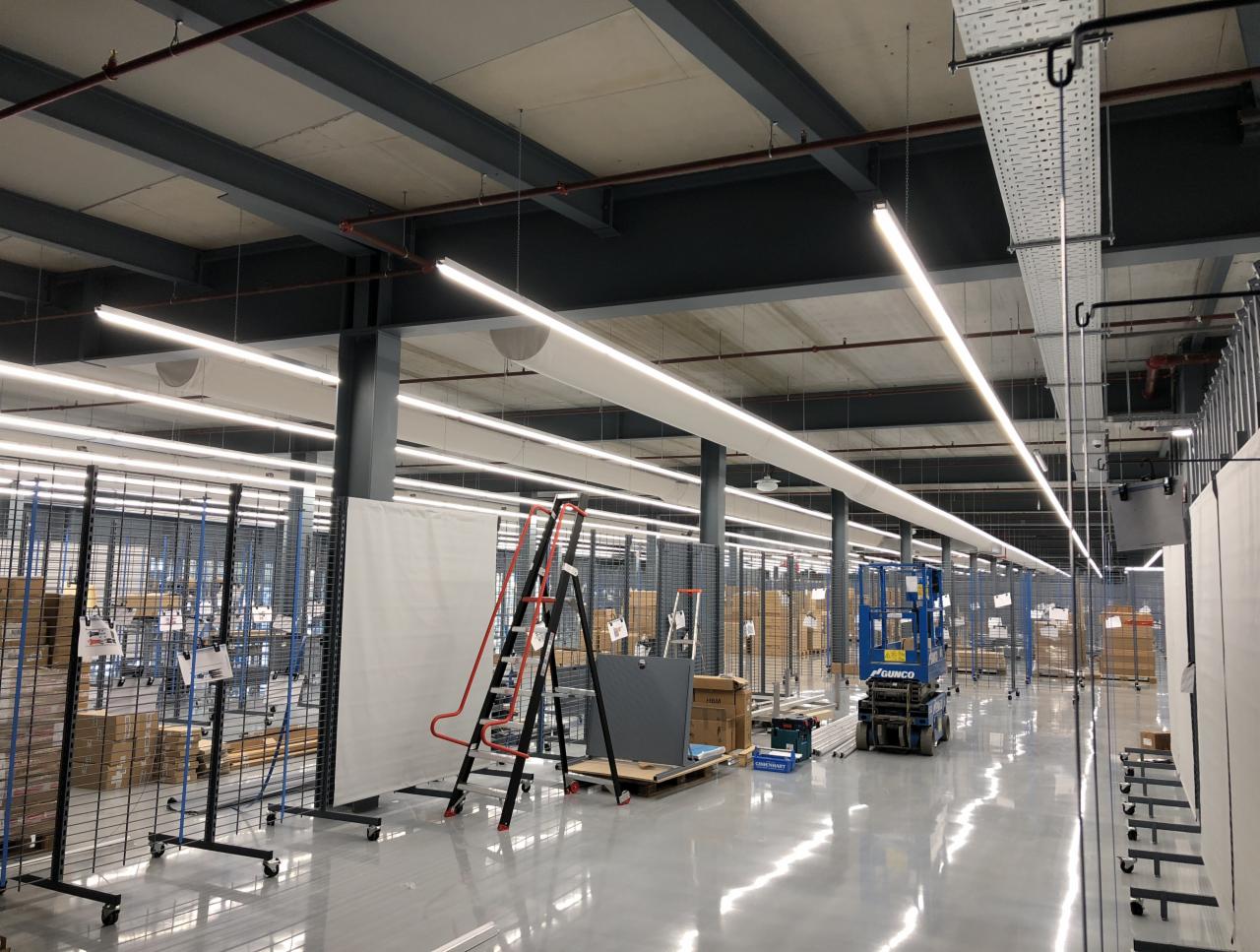 Montaż liniowego oświetlenia LED w hali magazynowej, widoczne drabiny, podnośnik i elementy konstrukcyjne sufitu.
