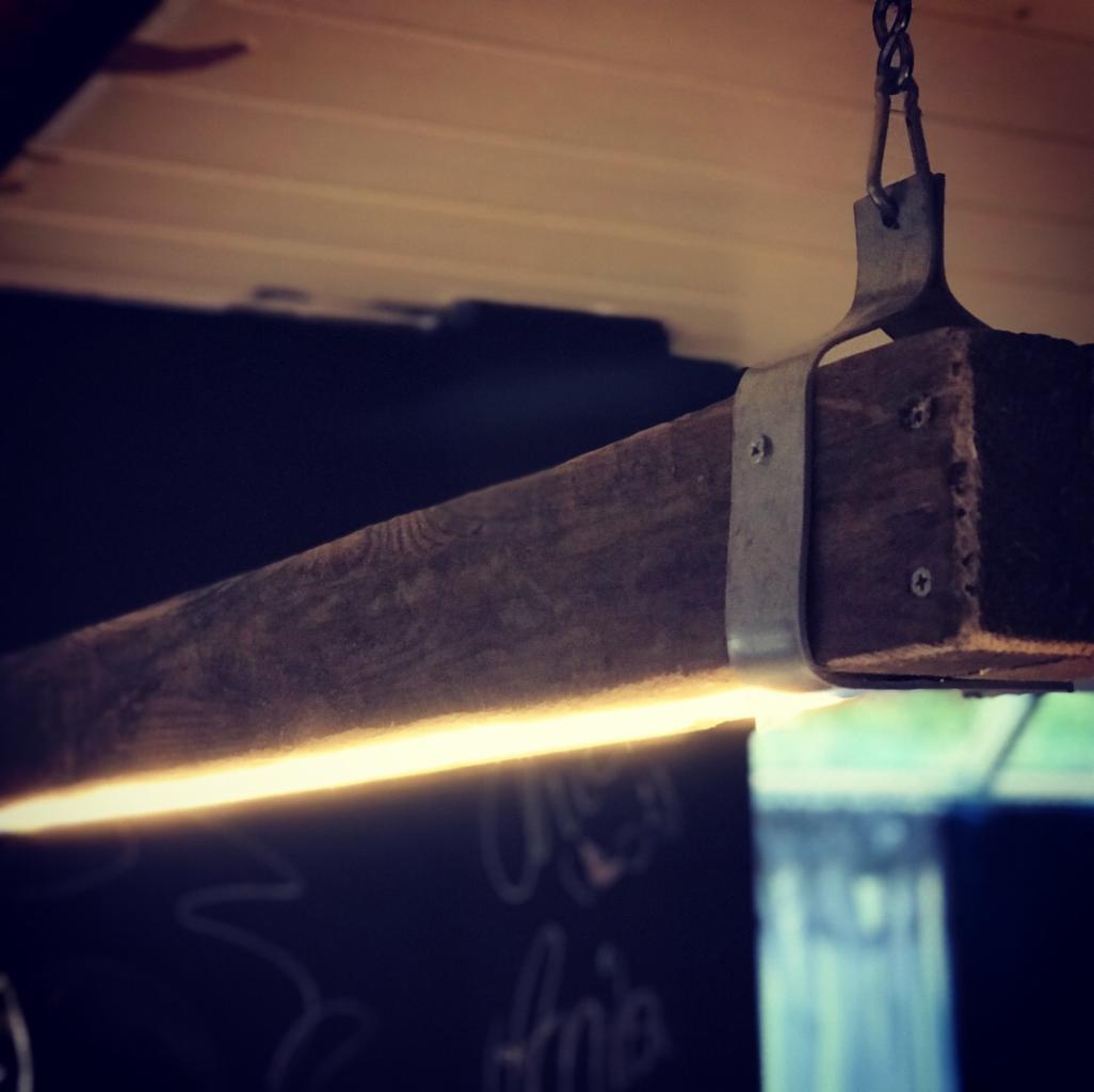 Drewniana belka z ukrytym źródłem światła LED, zawieszona na łańcuchu, mocowana metalową obejmą z widocznymi śrubami, na tle tablicy kredowej z napisem.