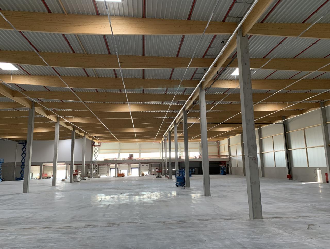 Wnętrze hali magazynowej w Sławnie, z widoczną konstrukcją drewnianą dachu i betonowymi słupami, przygotowywanej do instalacji oświetlenia.