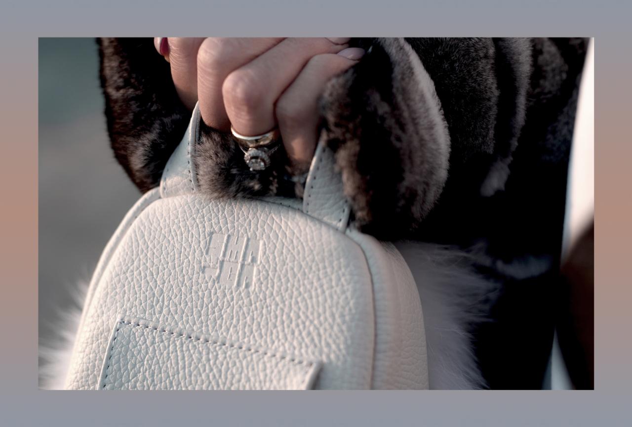 Kobieta w futrzanym okryciu trzyma biały skórzany plecak z wytłoczonym logo, eksponując pierścionek zaręczynowy.