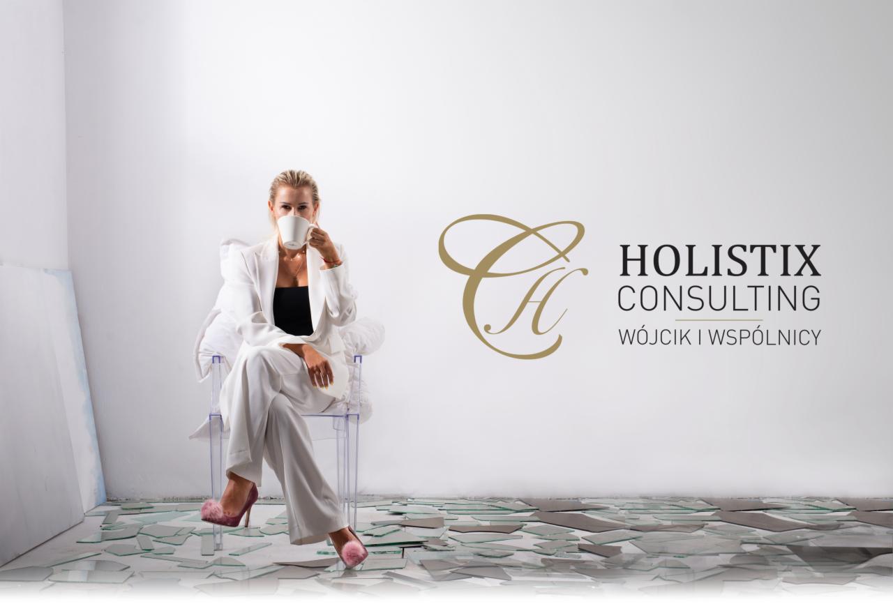 Kobieta w białym garniturze siedzi na krześle z filiżanką na tle ściany z logo firmy Holistix Consulting, podłoga pokryta rozbitym szkłem.