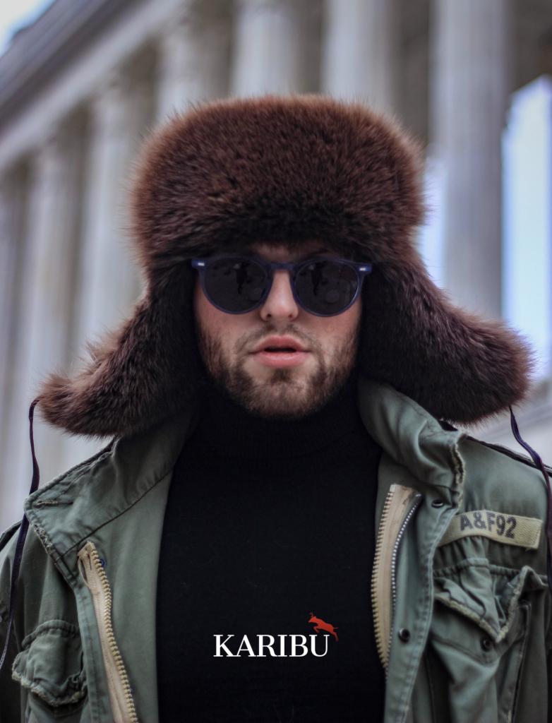 Mężczyzna z brodą w futrzanej czapce z klapkami, okularach przeciwsłonecznych i kurtce khaki z naszywką A&F92, na tle kolumn.
