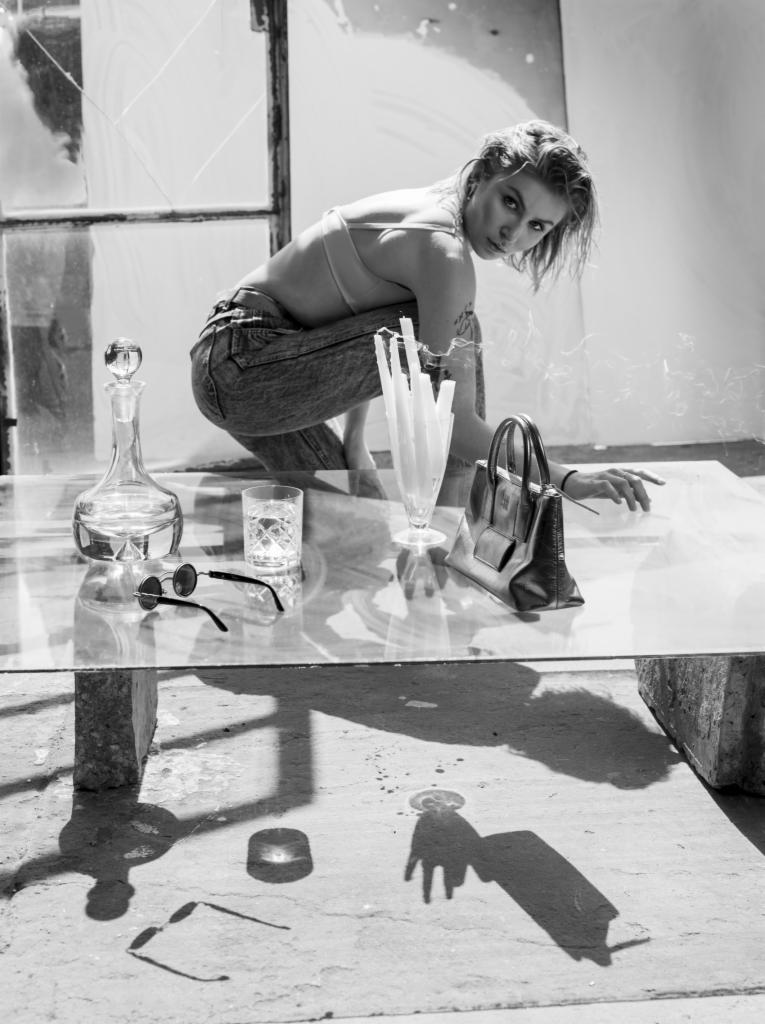 Czarno-białe zdjęcie kobiety w dżinsach kucającej za szklanym stołem, na którym znajdują się: karafka z wodą, szklanka, okulary przeciwsłoneczne, świece i torebka. W tle okno z rozbitymi szybami.