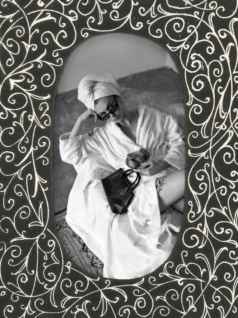 Portret kobiety w szlafroku i ręczniku na głowie, siedzącej na łóżku z talerzem ciastek i torebką, ujęty w ozdobną ramkę z motywami roślinnymi, czarno-biały.