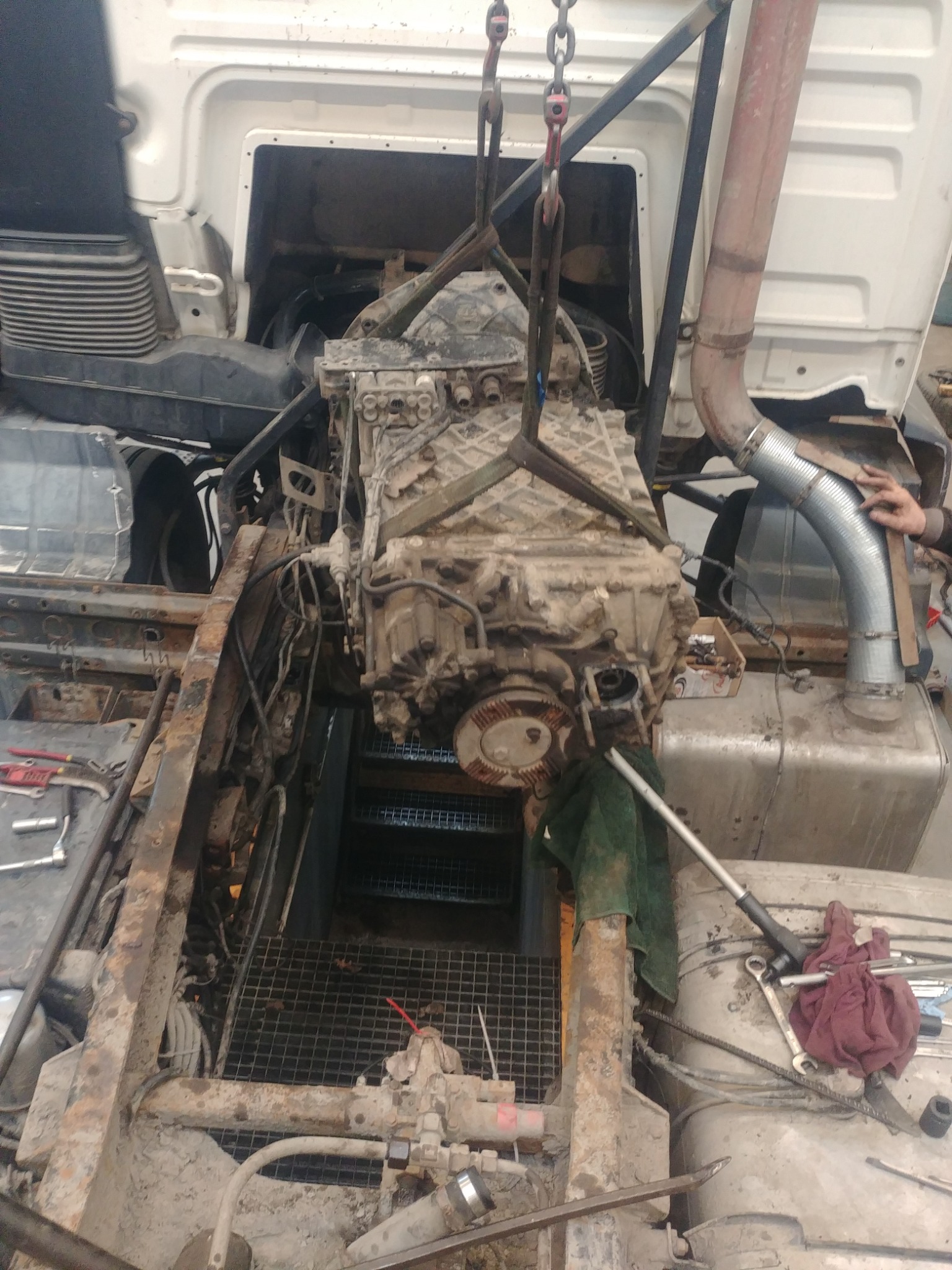 Brudny silnik ciężarówki zawieszony na łańcuchach podczas wymiany, widoczne narzędzia i elementy konstrukcyjne pojazdu.