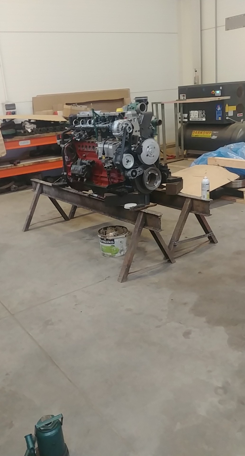 Silnik spalinowy na metalowym stojaku w warsztacie, widoczny pojemnik z farbą obok.