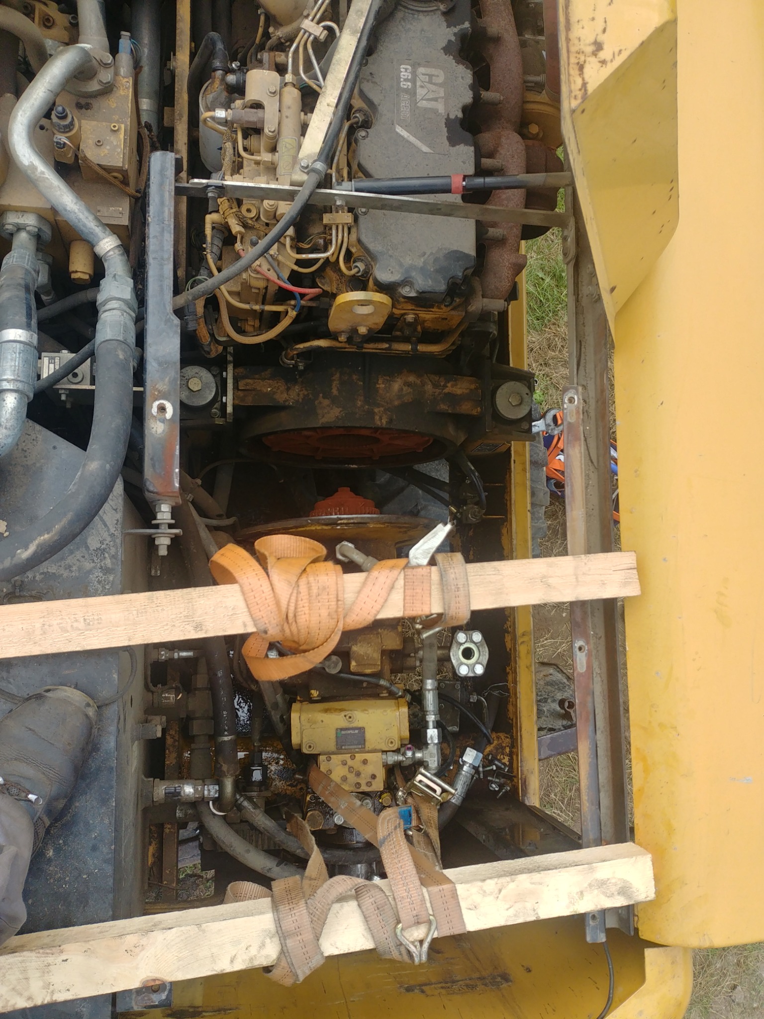 Widok z góry na silnik maszyny budowlanej Caterpillar, zabezpieczony drewnianymi belkami i pasami transportowymi, widoczne liczne przewody i elementy hydrauliczne, ślady oleju i zabrudzenia...