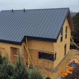 KRAUS-KAM SPÓŁKA Z OGRANICZONĄ ODPOWIEDZIALNOŚCIĄ - Surowy stan domu z drewnianą konstrukcją i szarym dachem z blachy na działce z ziemią, widoczne rusztowania i żółte rury doprowadzające media.