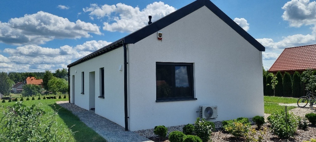 Nowoczesny, parterowy dom z białym tynkiem, czarnym dachem i oknami, klimatyzacja na ścianie, alarm, otoczony zielenią i kostką brukową, widok na błękitne niebo z obłokami.