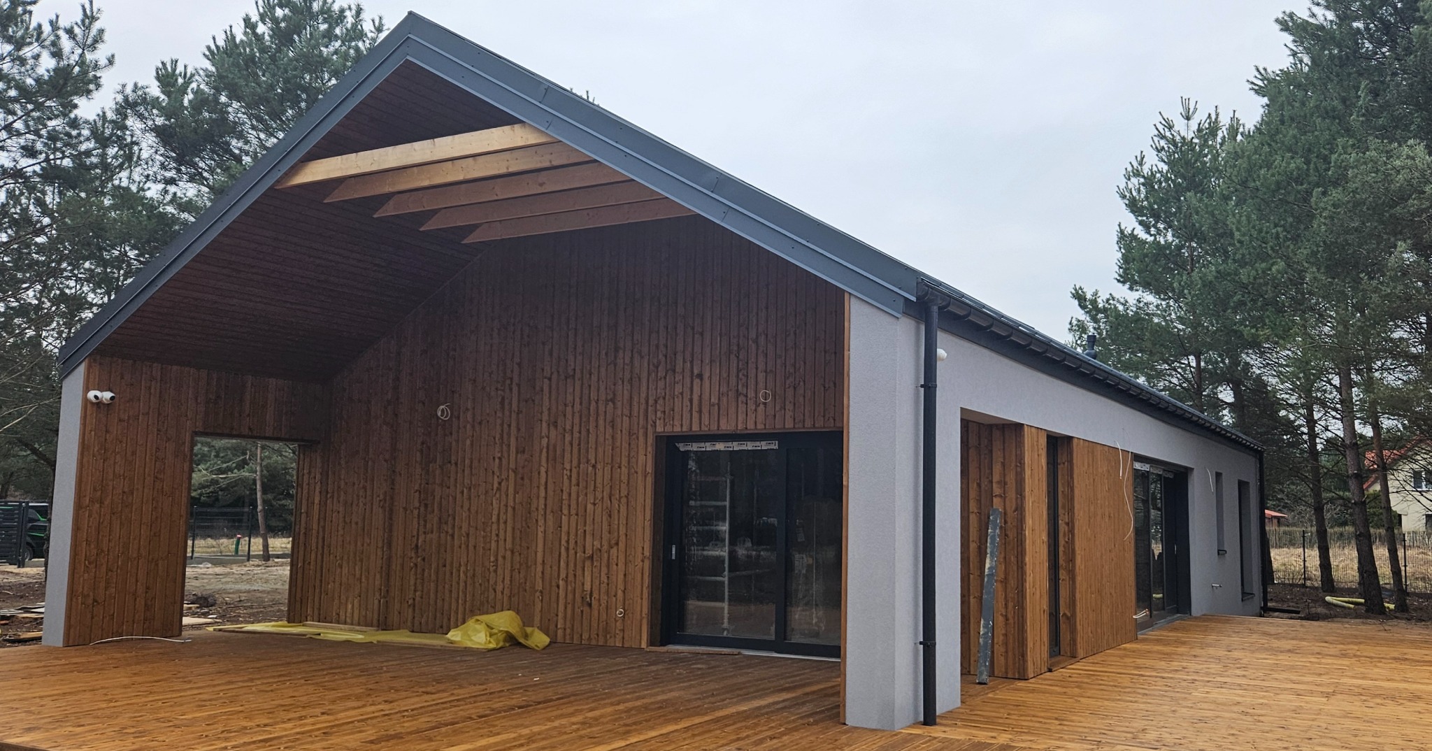 Nowoczesny budynek z drewnianą elewacją i dużym tarasem z desek, widoczne elementy wykończeniowe i otaczający las.