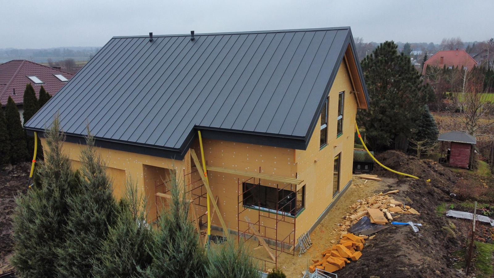 Surowy stan domu z drewnianą konstrukcją i szarym dachem z blachy na działce z ziemią, widoczne rusztowania i żółte rury doprowadzające media.