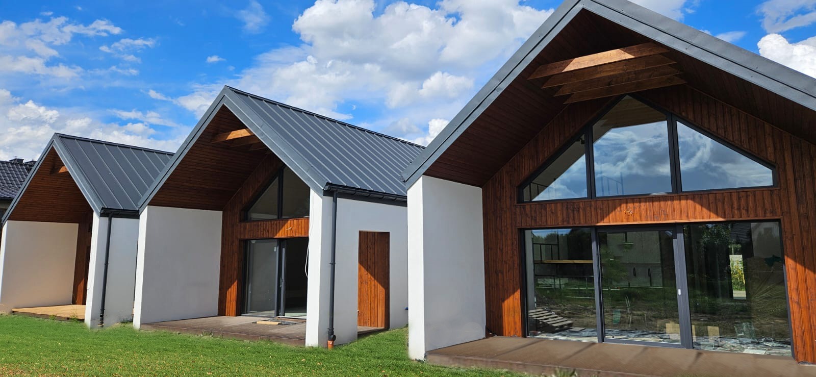 Trzy nowoczesne budynki z drewnianą elewacją i dużymi trójkątnymi oknami, pokryte grafitową blachą na tle błękitnego nieba z chmurami.