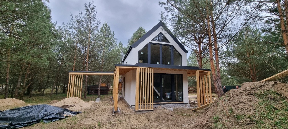 Nowoczesny dom z dużymi oknami i drewnianą konstrukcją werandy w otoczeniu sosnowego lasu, widoczny stos piasku i ziemi sugerujący trwające prace wykończeniowe.