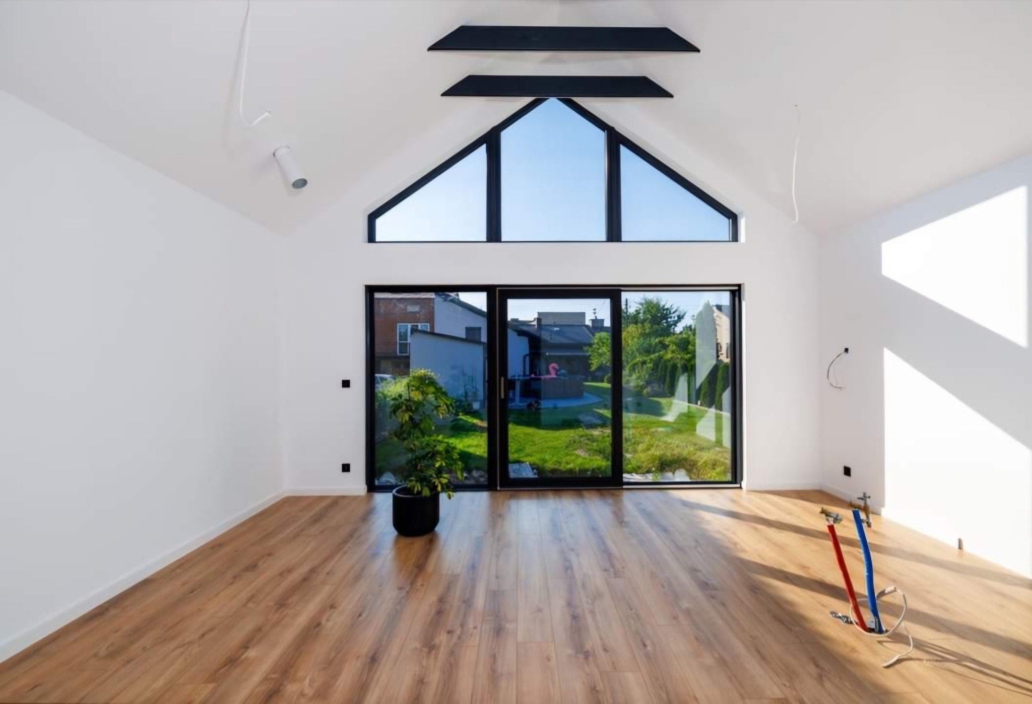 Wnętrze domu w stylu nowoczesnej stodoły z dużym oknem trójkątnym i przesuwnymi drzwiami tarasowymi, widoczne instalacje wodne w podłodze, parkiet i roślina doniczkowa.