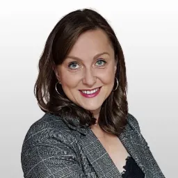 Portret kobiety w szarej marynarce w kratę i srebrnych kolczykach kołach, uśmiechającej się i patrzącej wprost, na jasnoszarym tle.