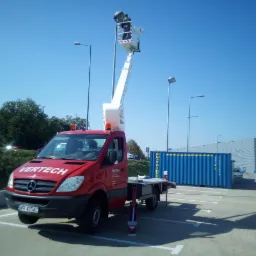 Czerwony samochód z podnośnikiem koszowym VERTECH na parkingu, pracownik w koszu na tle błękitnego nieba i kontenera.