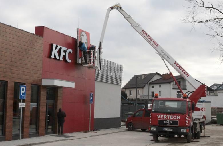 Montaż logo KFC na budynku restauracji przy użyciu podnośnika koszowego Vertech. Pracownik w koszu na wysokości, drugi mężczyzna na dole obserwuje. W tle budynki mieszkalne.