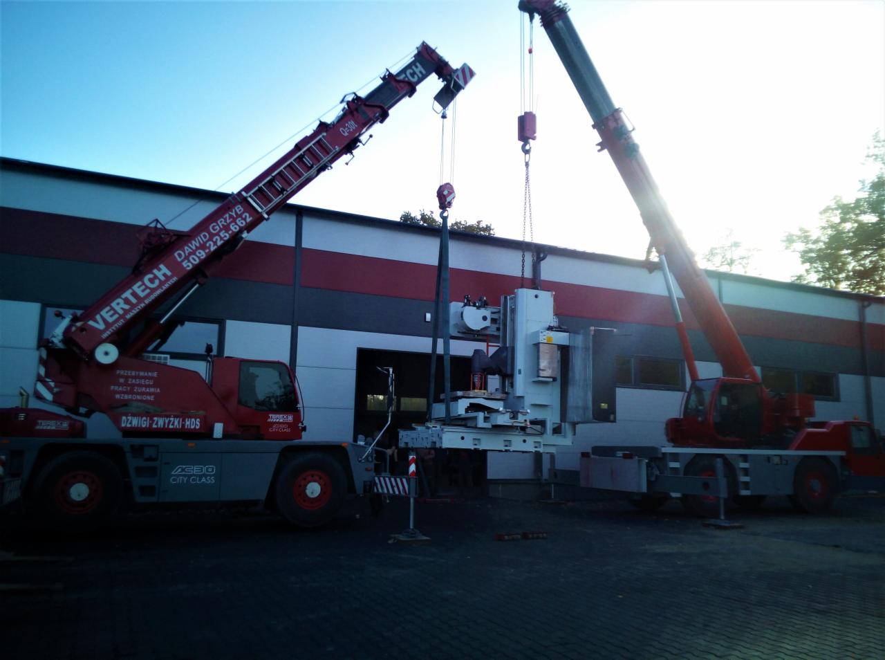 Dwa czerwone dźwigi Terex AC30 City Class podnoszą ciężką maszynę na tle szaro-białego budynku z czerwonym pasem, widoczne liny i haki dźwigowe.