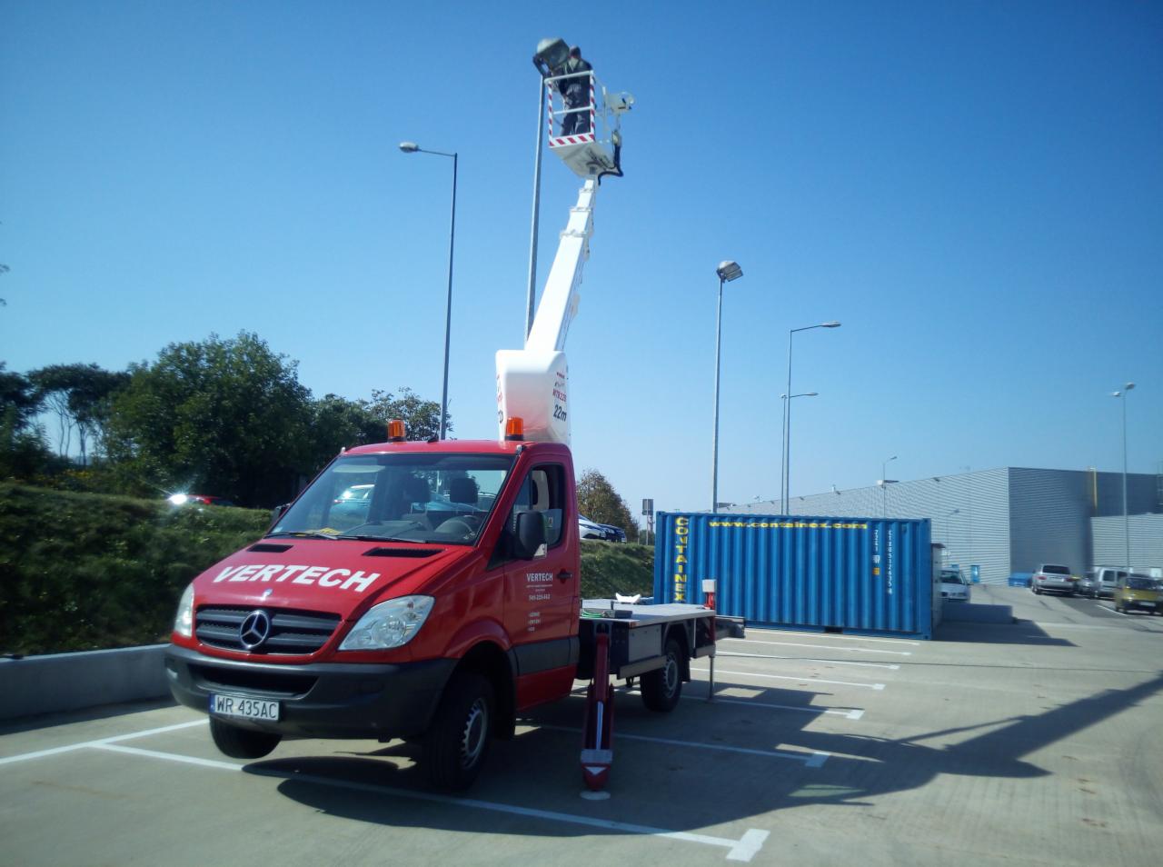Czerwony samochód z podnośnikiem koszowym VERTECH na parkingu, pracownik w koszu na tle błękitnego nieba i kontenera.