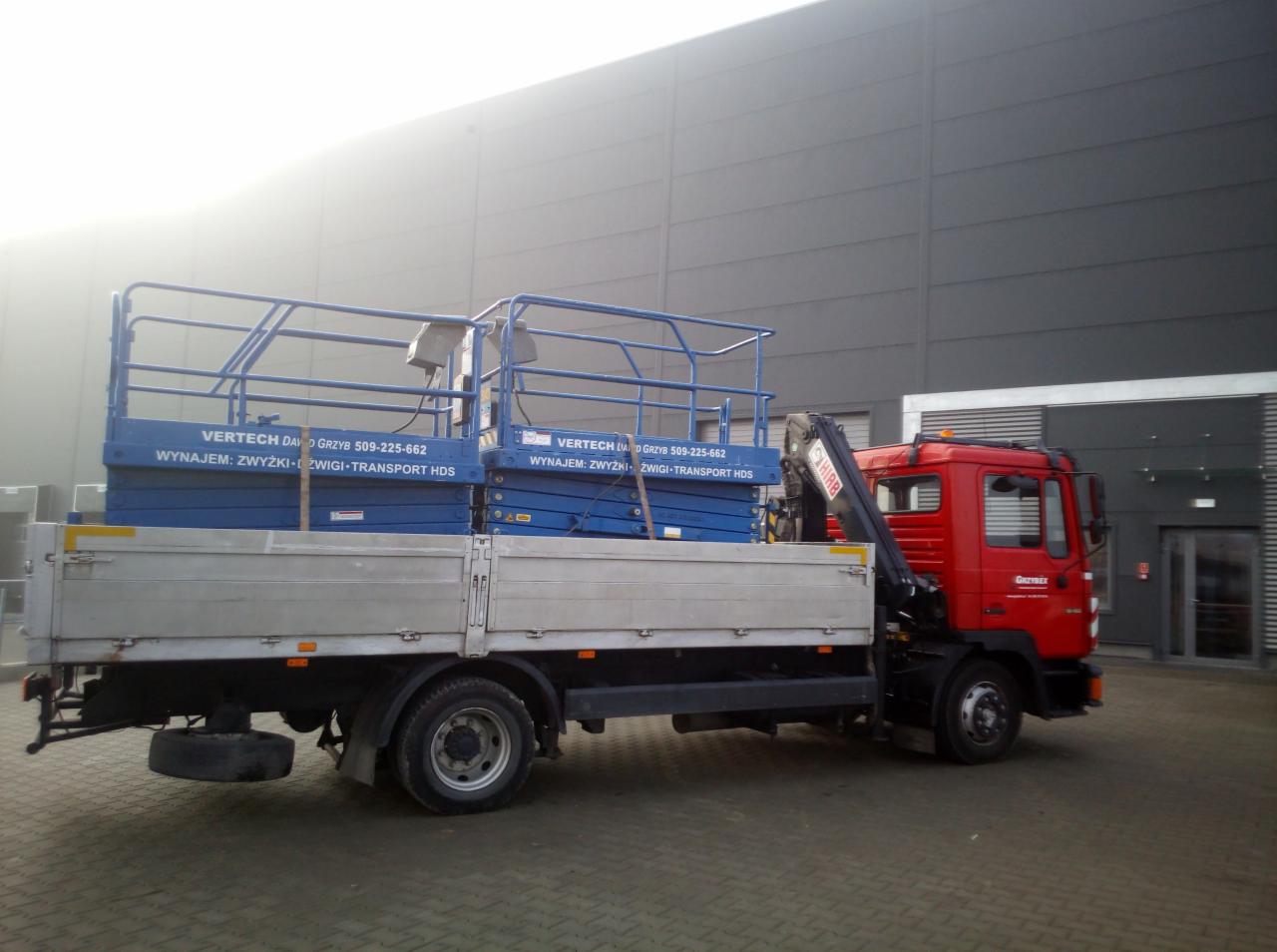 Ciężarówka z czerwoną kabiną przewożąca dwie niebieskie platformy robocze na tle szarego budynku przemysłowego. Na platformach widoczne napisy 'VERTECH DAVID GRZYB 509-225-662 WYNAJEM...