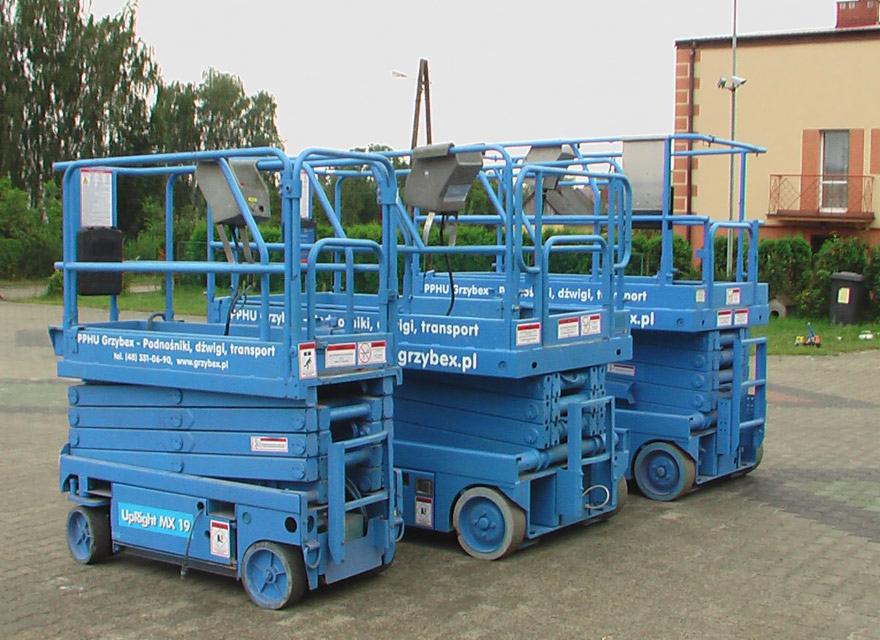 Trzy niebieskie podnośniki nożycowe UpRight MX 19 zaparkowane obok siebie, z widocznymi detalami konstrukcyjnymi i oznaczeniami firmy Grzybex.