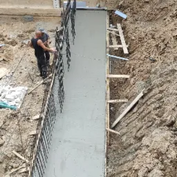 Wylewanie betonu na fundament z zamontowanym zbrojeniem pionowym. Widoczni dwaj pracownicy w trakcie pracy.