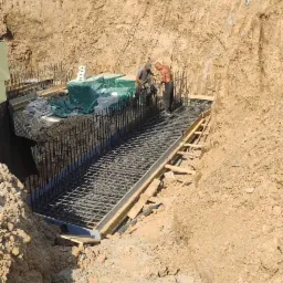 Dwóch robotników podczas wiązania zbrojenia w wykopie pod fundament, widoczne drewniane szalunki i izolacja pionowa.