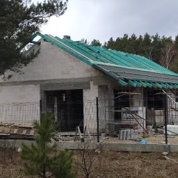 OPTYplus BUD Sp. z o.o. - Surowy stan domu jednorodzinnego z bloczków, z widoczną konstrukcją dachu w kolorze zielonym, otoczonego siatką ogrodzeniową i drzewami.