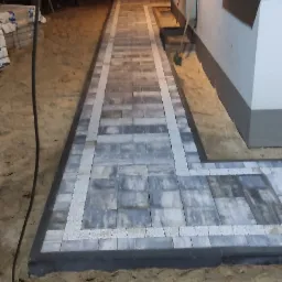 Ułożona ścieżka z szarej kostki brukowej z jasnymi obrzeżami, oświetlona lampami ściennymi, widoczna w nocy na piaszczystym podłożu.