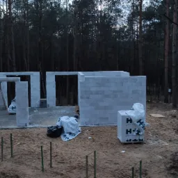 Wznoszenie ścian parterowego domu z bloczk&oacute;w betonowych na fundamencie, widok z g&oacute;ry na tle sosnowego lasu, widoczne palety z materiałem budowlanym i zbrojenie w ziemi.