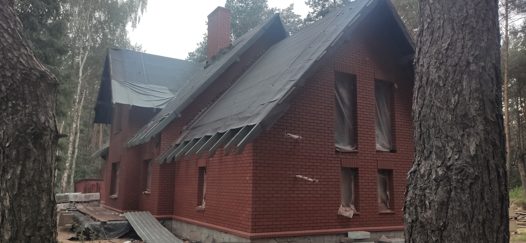Niedokończony dom murowany z czerwonej cegły, z oknami zabezpieczonymi folią, widok z zewnątrz na tle drzew, dach częściowo pokryty czarną folią ochronną, w tle widoczny komin z cegły.