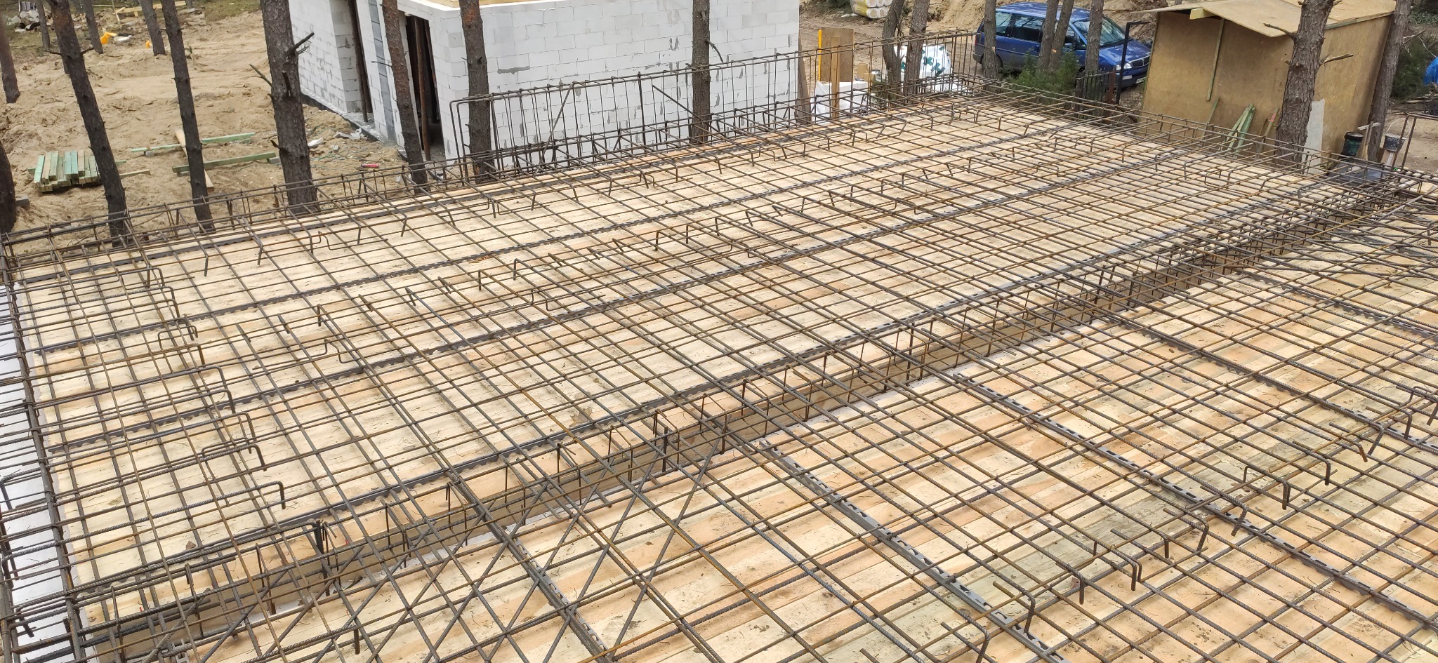 Siatka zbrojeniowa na deskowaniu przygotowana do zalania betonem, widoczny budynek w stanie surowym otwartym w tle