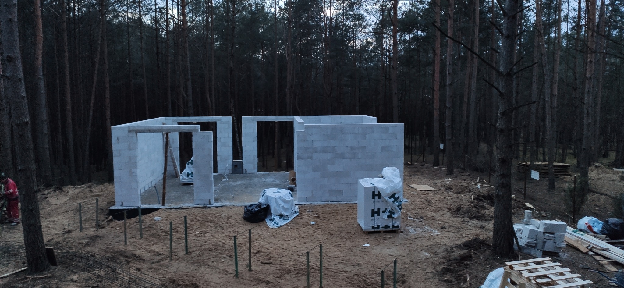 Wznoszenie ścian parterowego domu z bloczków betonowych na fundamencie, widok z góry na tle sosnowego lasu, widoczne palety z materiałem budowlanym i zbrojenie w ziemi.