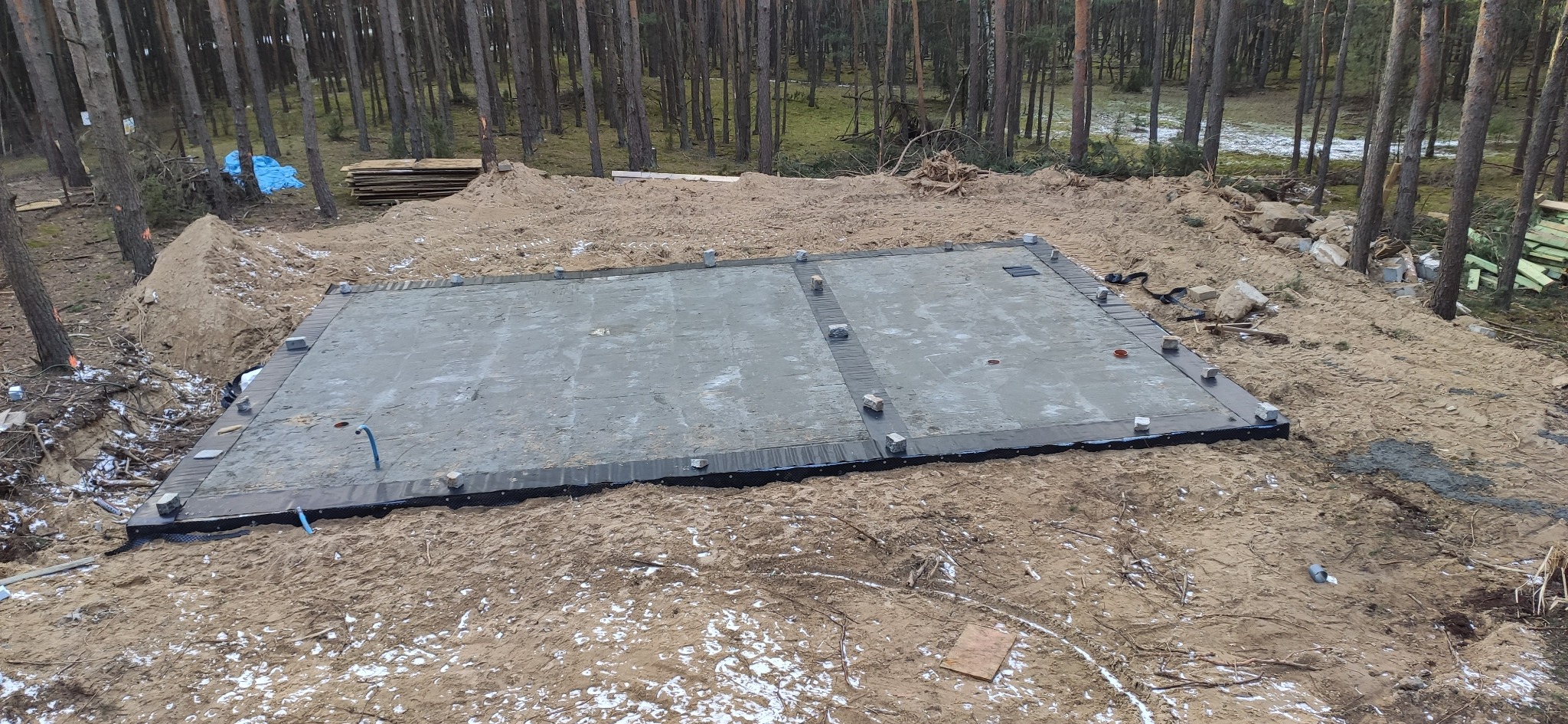 Zalana płyta fundamentowa w lesie, zabezpieczona czarną folią, z widocznymi wystającymi rurami instalacyjnymi i elementami szalunku.