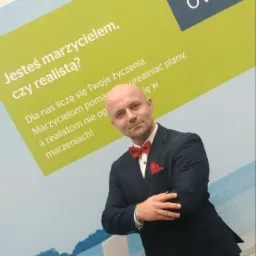 Mężczyzna w garniturze z czerwoną muchą i poszetką, pozujący na tle baneru OVB z hasłem 'Jesteś marzycielem, czy realistą?'