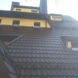 Widok z dołu na dom z brązowym dachem z dachówki, żółtą elewacją i ciemnymi rynnami, z widoczną częścią komina.