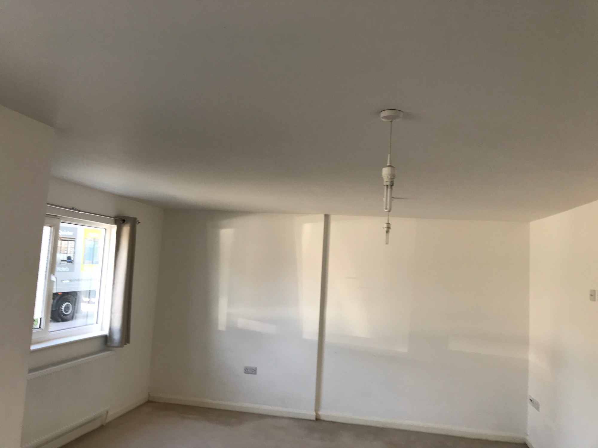 Pomalowane na biało wnętrze pokoju z grzejnikiem pod oknem, widoczne gniazdka elektryczne i wisząca lampa bez klosza.